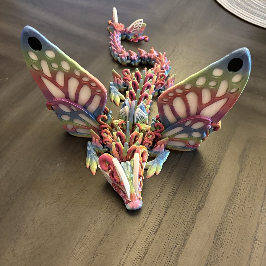 Articulated butterfly-wing dragon 3D printed in a rainbow gradient, posed on a wood table
